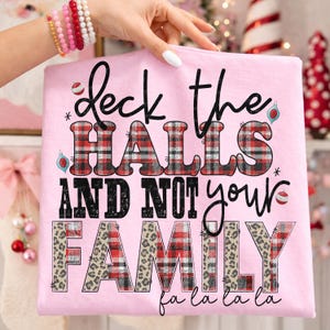 Puede incluir: Camiseta rosa claro con el texto "Deck the HALLS AND NOT your FAMILY" en negro, cuadros rojos y estampado de leopardo. La frase "fa la la la la" está en la parte inferior. Ropa temática navideña.