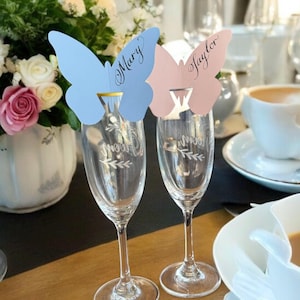 May include: Two champagne flutes with butterfly-shaped place cards. One card is light blue with the name "Mary" and the other is pink with the name "Taylor". The flutes are on a wooden table, with a floral arrangement and a cup of coffee in the background.