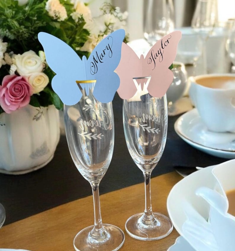 May include: Two champagne flutes with butterfly-shaped place cards. One card is light blue with the name "Mary" and the other is pink with the name "Taylor". The flutes are on a wooden table, with a floral arrangement and a cup of coffee in the background.