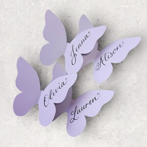 May include: Four lavender butterfly-shaped place cards. Each butterfly has a name written in elegant script: Jenna, Alison, Olivia, and Lauren. The place cards are arranged on a light gray surface, perfect for a wedding or special event.