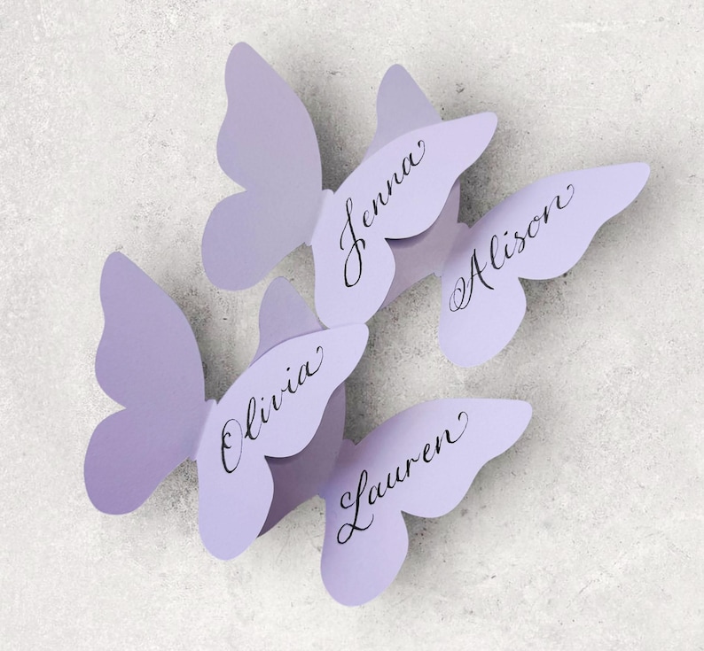 May include: Four lavender butterfly-shaped place cards. Each butterfly has a name written in elegant script: Jenna, Alison, Olivia, and Lauren. The place cards are arranged on a light gray surface, perfect for a wedding or special event.