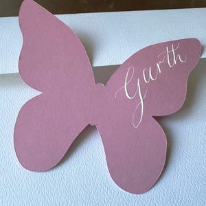 May include: A mauve butterfly-shaped place card with the name "Garth" in elegant white script. The card is made of paper and is likely used for a wedding or event.