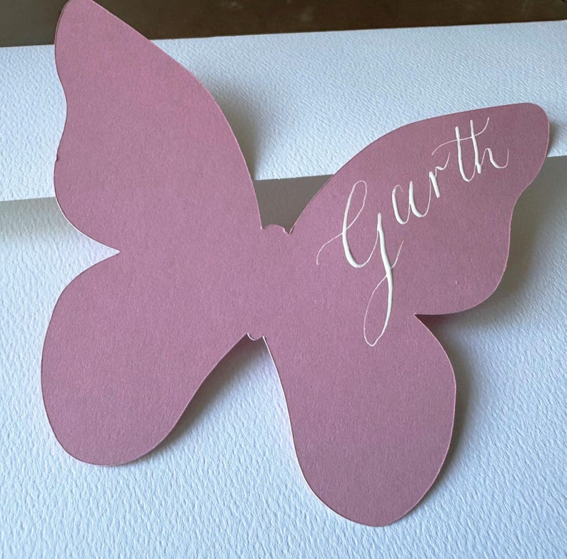 May include: A mauve butterfly-shaped place card with the name "Garth" in elegant white script. The card is made of paper and is likely used for a wedding or event.