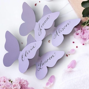 May include: Four lavender butterfly-shaped place cards with names written in elegant script: Jenna, Alison, Olivia, and Lauren. The cards are arranged on a white surface, with pink flower petals and blooms scattered around.