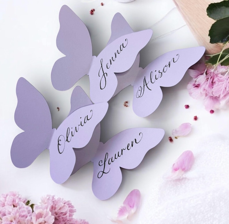 May include: Four lavender butterfly-shaped place cards with names written in elegant script: Jenna, Alison, Olivia, and Lauren. The cards are arranged on a white surface, with pink flower petals and blooms scattered around.
