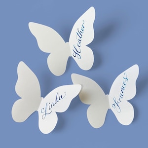 May include: Three white butterfly-shaped place cards with names written in blue script: "Heather", "Linda", and "Frances". The cards are arranged on a light blue background, ideal for weddings or events.