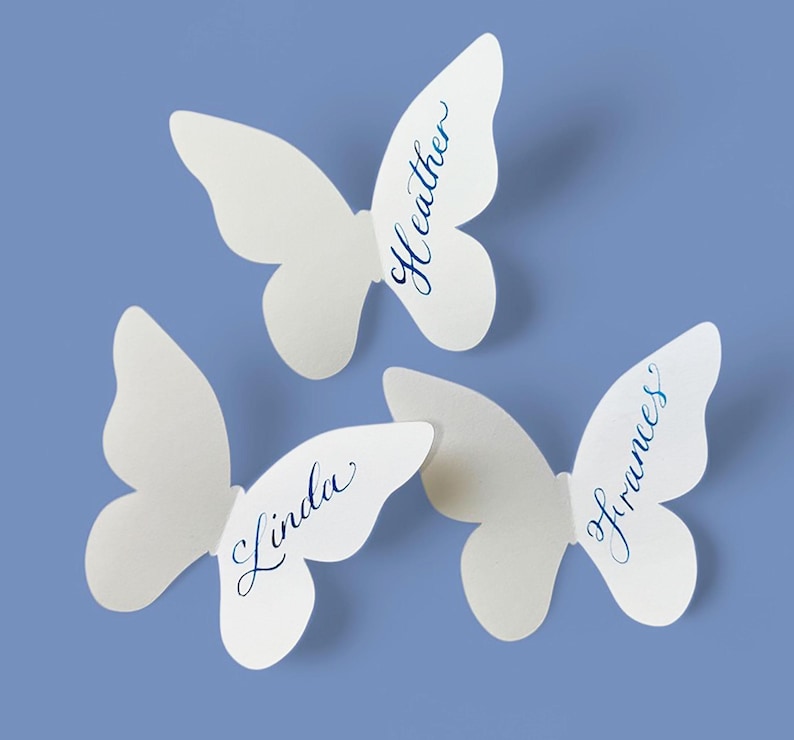 May include: Three white butterfly-shaped place cards with names written in blue script: "Heather", "Linda", and "Frances". The cards are arranged on a light blue background, ideal for weddings or events.