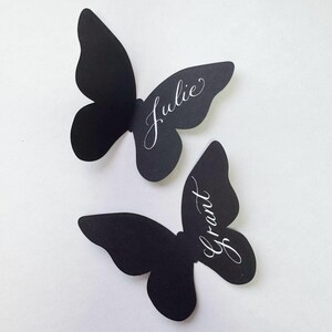 May include: Two black butterfly-shaped place cards. The name "Julie" is written in elegant white script on the upper butterfly, and "Grant" is written on the lower butterfly. The place cards are likely used for weddings or special events.