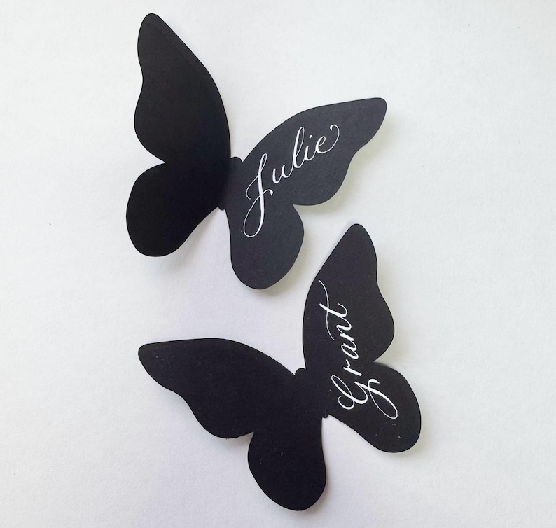 May include: Two black butterfly-shaped place cards. The name "Julie" is written in elegant white script on the upper butterfly, and "Grant" is written on the lower butterfly. The place cards are likely used for weddings or special events.
