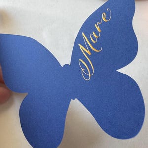 May include: A blue butterfly-shaped paper cutout with gold script text that reads "Mare". The butterfly has a symmetrical design with rounded wings and a textured surface. The background is white.