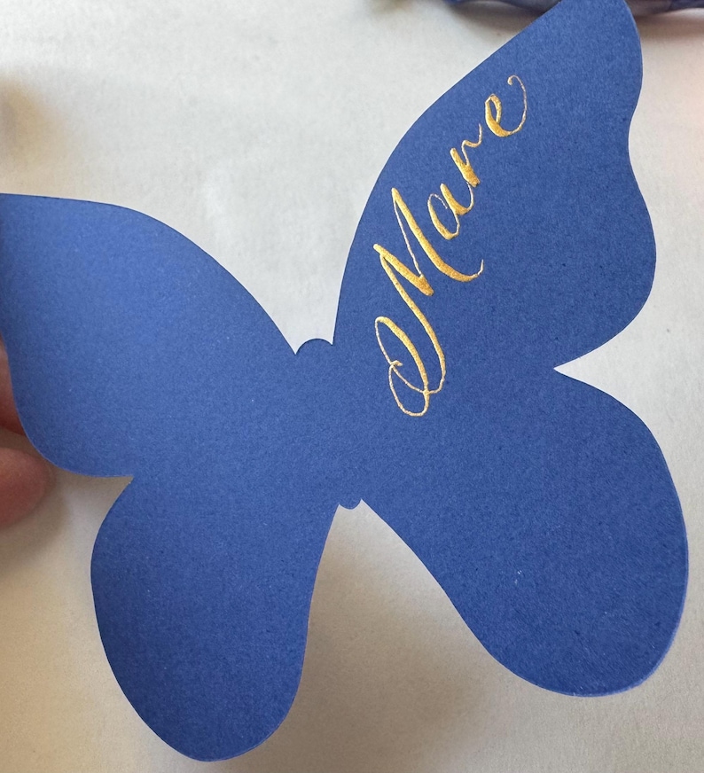 May include: A blue butterfly-shaped paper cutout with gold script text that reads "Mare". The butterfly has a symmetrical design with rounded wings and a textured surface. The background is white.