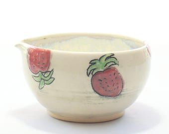 May include: Cream-colored ceramic bowl with a spout and hand-painted strawberries. The bowl features two red strawberries with green leaves, adding a touch of whimsy. The bowl is ideal for serving or mixing ingredients.