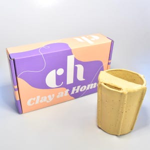 Könnte beinhalten: Ein gelber Keramik-Tontopf mit strukturierter Oberfläche und rechteckiger Öffnung. Daneben eine Box mit dem Aufdruck "ch" und "Clay at Home". Die Box hat ein lila-pfirsichfarbenes Farbschema.