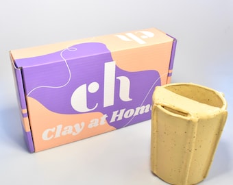 May include: A yellow ceramic clay pot with a textured surface and a rectangular opening. Beside it is a box with the text "ch" and "Clay at Home" printed on it. The box has a purple and peach color scheme.
