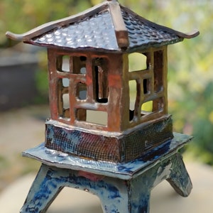 Könnte beinhalten: Eine Keramiklaterne im Pagodenstil mit dunkelblauer, brauner und kupferfarbener Oberfläche. Die Laterne hat ein mehrstöckiges Dach und einen quadratischen Körper mit offenen Seiten. Der Sockel ist dunkelblau mit einem strukturierten Muster. Die Laterne ist etwa 25 cm hoch.