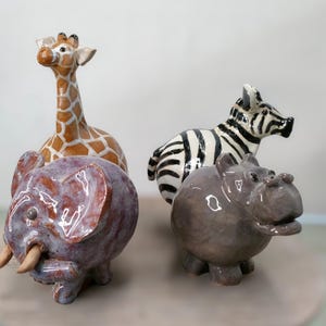 May include: A collection of four glazed ceramic animal figurines including a tall giraffe with brown and white patches, a black and white striped zebra, a round purple elephant with two tusks, and a stout grey hippopotamus with a wide mouth.