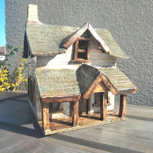 May include: A ceramic house sculpture featuring white walls, a textured green shingled roof, and a brown wooden-style porch with supporting pillars. The structure includes a chimney, a front-facing dormer window, and a detailed porch floor.