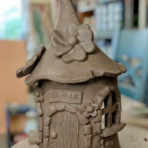 May include: A handcrafted clay fairy house with a whimsical design. The house features a curved roof, a door, and a window, all adorned with floral accents. The sign above the door reads "THE HOLE".