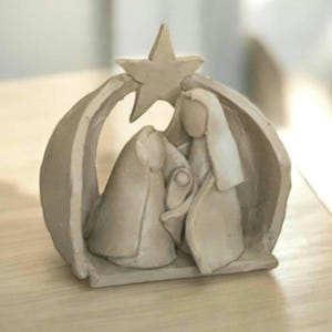 Puede incluir: Una escultura de belén beige. La escultura representa a María, José y el niño Jesús, resguardados bajo un arco con una estrella en la parte superior. Las figuras son estilizadas y el diseño general es sencillo.