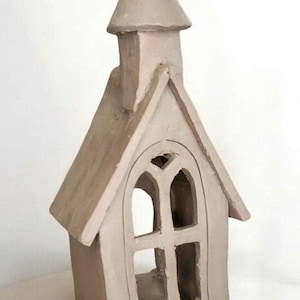 May include: A light beige, unglazed ceramic sculpture shaped like a small church or chapel. The structure features a pointed roof, a tall steeple, and an arched window frame with a central mullion, creating an open, hollow interior space.