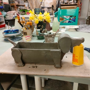 May include: A gray clay dachshund planter with a brown interior. The planter is sitting on a wooden table with a yellow container next to it.