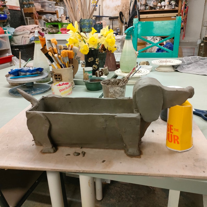 May include: A gray clay dachshund planter with a brown interior. The planter is sitting on a wooden table with a yellow container next to it.