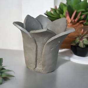 May include: A grey concrete flower pot shaped like a tulip. The pot has six petals and is suitable for small plants. The pot is on a white surface, with other plants in the background.