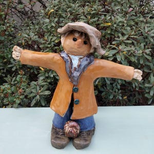 May include: A ceramic figurine of a scarecrow wearing a tan coat with two green buttons, a grey scarf, and a floppy hat. The figure has blue trousers, dark brown shoes, and a small pumpkin at its feet, with arms outstretched to the sides.