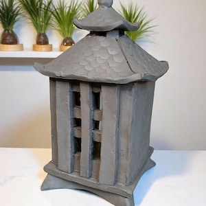 May include: A gray ceramic pagoda-style lantern with a textured roof and vertical bars. The lantern has a square base and a small, rounded finial on top. The design is reminiscent of traditional Japanese garden decor.