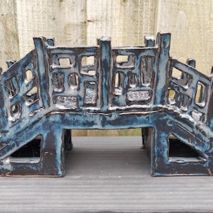 May include: A ceramic bridge sculpture featuring a dark charcoal base with a glossy blue glaze finish. The structure has an arched design with open rectangular cutouts along the railings and two large triangular openings at the base.