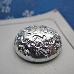 May include: A silver-colored, domed button with an ornate, raised floral design. The button is round and has a shiny, metallic finish. The button is likely for sewing or crafting projects.