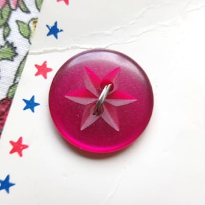May include: A circular, translucent magenta button featuring a six-pointed star design embossed in the center. The button is attached with a silver-colored metal wire loop and is positioned on a white card decorated with small red and blue stars.