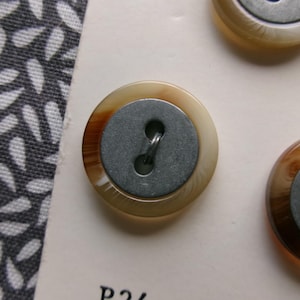 May include: Close-up of several round buttons with a light brown and dark brown marbled effect. Each button has a gray metal center with two holes for sewing. The buttons are on a cream-colored surface next to a patterned fabric.