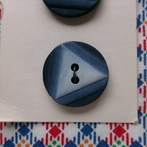 May include: Two navy blue buttons with a triangular design on a cream-colored card. The buttons have two holes for sewing. The background is a colorful fabric with a geometric pattern in red, blue, and white.