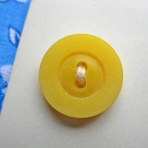 Könnte beinhalten: Nahaufnahme eines leuchtend gelben Knopfes mit einem Vier-Loch-Design und einem weißen Faden. Der Knopf ist rund mit einer leicht erhöhten Mitte und einer glatten, matten Oberfläche. Der Knopf liegt auf einer weißen Oberfläche.