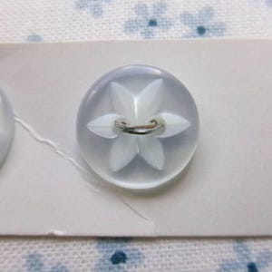 May include: Two round, translucent, light blue buttons attached to a white card. Each button features a raised, white, five-petaled floral design in the center with a metal shank on the back. The card has the text "SIZE" and "COUNT" printed on it.