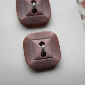 May include: Two square-shaped buttons with a light pink color and a silver metal shank. The buttons have a raised, textured design.