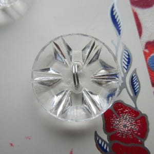Könnte beinhalten: Klarer, facettierter Glas-Knopf mit Sternmuster. Der Knopf hat eine Schlaufe auf der Rückseite zum Annähen. Der Knopf liegt auf einer weißen Oberfläche mit floral gemustertem Papier.