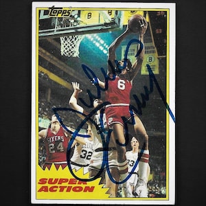 May include: A vintage Topps Super Action basketball trading card featuring a basketball player in a red and white uniform with the number 6 on the front. The player is jumping up to shoot the ball. The card has a blue signature on the front. The card is in a white border with a yellow background.