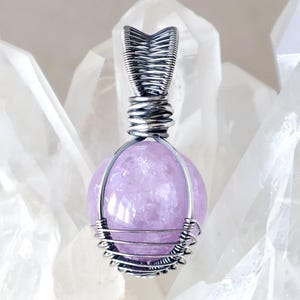 May include: A pendant with a lavender gemstone, encased in silver-coloured wire. The wire is carefully wrapped around the spherical stone and forms a decorative bail. The pendant is set against a backdrop of clear quartz crystals.