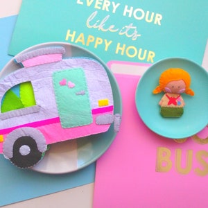 Peut inclure: Un camping-car en feutre et une poupée en feutre sur des assiettes turquoise, avec une carte turquoise qui dit "EVERY HOUR like it's HAPPY HOUR" et une carte rose qui dit "BUSY". Le camping-car est rose, gris et turquoise. La poupée a les cheveux orange et une croix rouge.