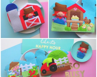 May include: A collection of colorful felt farm-themed decorations. The set includes a red barn, a silo, a chicken coop with chickens, a farmer tending a garden, and a tractor. The decorations are placed on plates with cards that say "Happy Hour" and "So Busy".