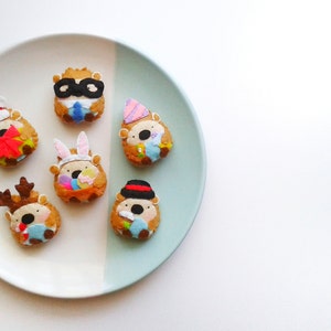 May include: A plate displaying seven hedgehog-shaped biscuits, each adorned with various festive decorations. One has a Santa hat, another a superhero mask, and others feature bunny ears, a party hat, a deer antler, and a bowler hat. The biscuits are on a two-tone plate.