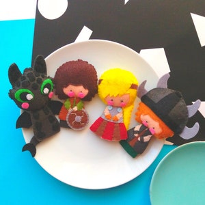 May include: A collection of handmade felt figures, inspired by "How to Train Your Dragon", are displayed on a white plate. The characters include a black dragon and three Vikings, each with unique outfits and accessories.