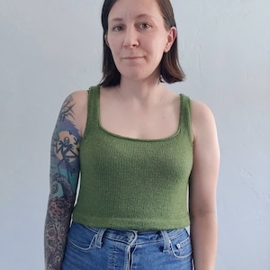 Könnte beinhalten: Eine Frau trägt ein grünes Stricktop mit quadratischem Ausschnitt. Sie trägt eine blaue Jeans und hat ein Tattoo auf ihrem Arm.