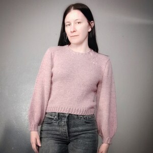 May include: A pink knitted sweater with long sleeves and a round neckline. The sweater has a slightly cropped fit and a ribbed hem.