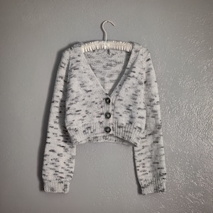 Könnte beinhalten: Ein weißer und grauer, kurzer Cardigan mit V-Ausschnitt und Knopfleiste. Der Cardigan hat lange Ärmel und ein gesprenkeltes Muster.