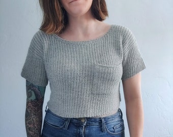 May include: A gray cropped sweater with a pocket on the front. The sweater is made of a soft, knit fabric. The model is wearing blue jeans.