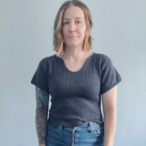 Puede incluir: Una mujer con un top de punto gris oscuro de manga corta y cuello en V. Lleva pantalones vaqueros azules y tiene un tatuaje en el brazo.