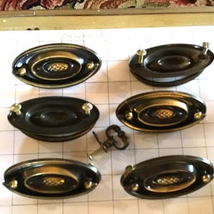 May include: Six oval drawer pulls with a dark bronze finish and gold accents. Each pull has a central decorative element and is mounted with small screws. A small key is also present.